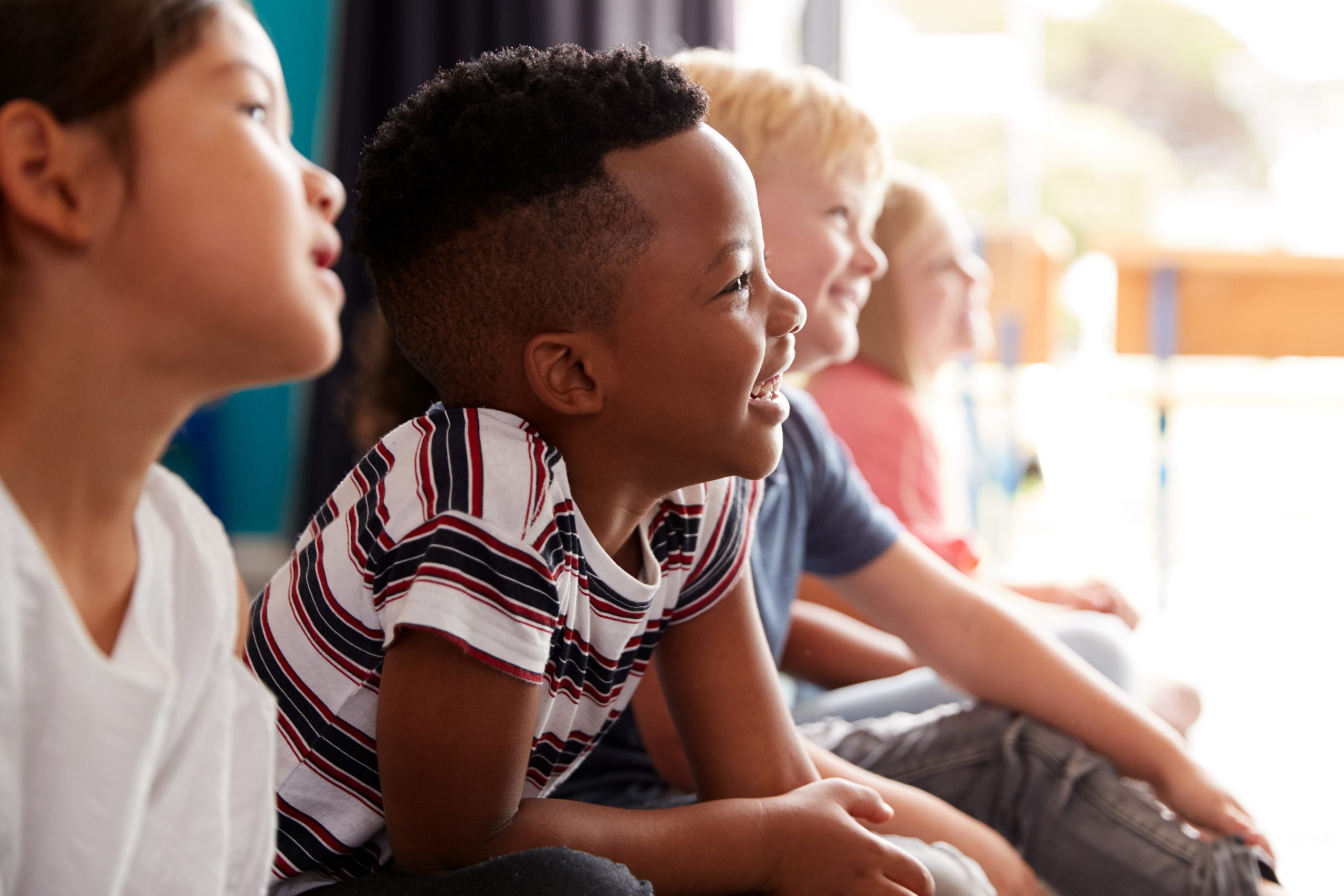 Group Of Elementary School Pupils Sitting On Floor Listening To Teacher Jenny Pentecost